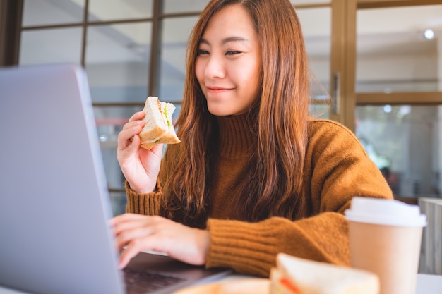 An asian woman holding and eating whole wheat sandwich while working on laptop computer