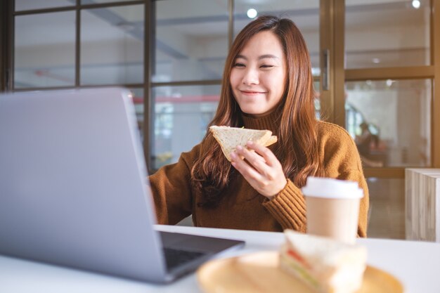 An asian woman holding and eating whole wheat sandwich while working on laptop computer