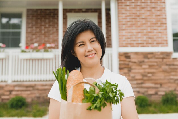 Photo asian woman holding a grocery bag with vegetables near her house