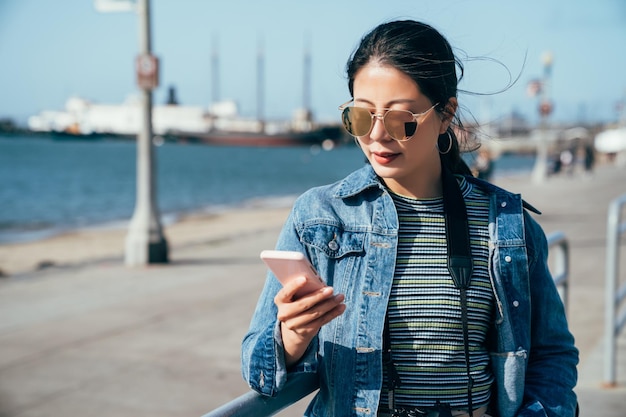 asian woman holding mobile phone searching online map app finding direction of hotel standing in the pier 39 next to clean blue ocean. lady in sunglasses looking at cellphone while sightseeing tour