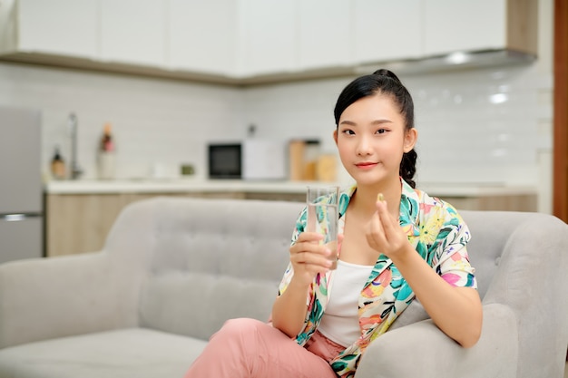 Asian woman holding pill and a glass of water sitting on sofa.