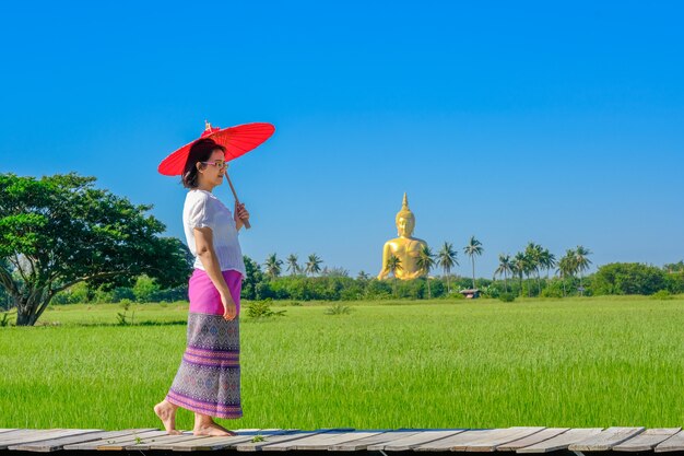 An Asian woman holding a red umbrella walking on a wooden bridge in the rice field with a large golden Buddha image.