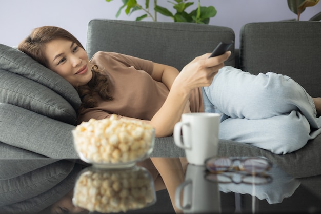 Donna asiatica che tiene il telecomando e guarda la tv con felicità mentre è sdraiata su un comodo divano con popcorn e un bicchiere di bevanda nel soggiorno di casa.