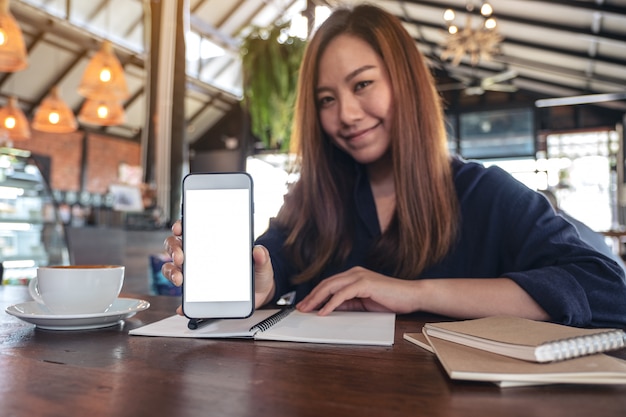 カフェでヴィンテージの木製のテーブルにコーヒーカップとノートブックと空白のデスクトップ画面で白い携帯電話を保持し、示すアジアの女性