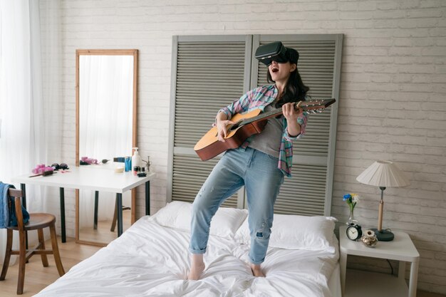 Азиатская женщина, держащая деревянную гитару, откинулась назад, чтобы сыграть на устройстве виртуальной реальности на своей кровати. Она кричит и вопит, как ее личный концерт.