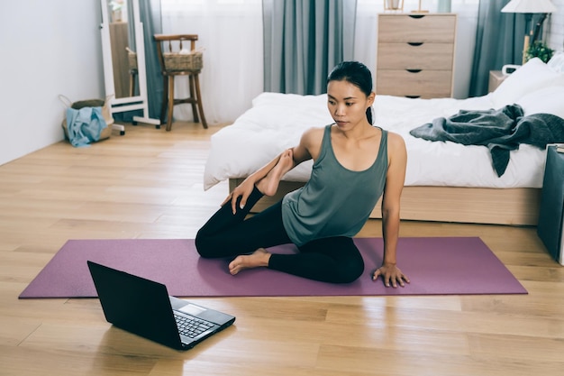 Asian woman under home isolation is taking online exercise\
course at home. taiwanese girl with dark skin is watching pc screen\
and listening to her coach’s instruction attentively