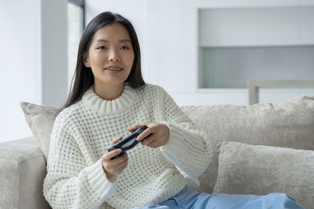 An asian woman is sitting on the couch holding a joystick and playing a game on the console the