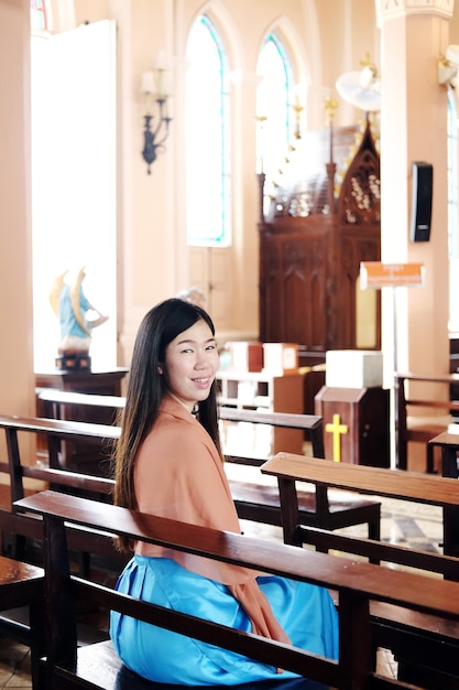 Asian woman is sitting on wooden chair and smiling for Pray to god in a Christian church for make a wish for hope and encouragement in life. Rituals and beliefs in Christian of Thailand concept.