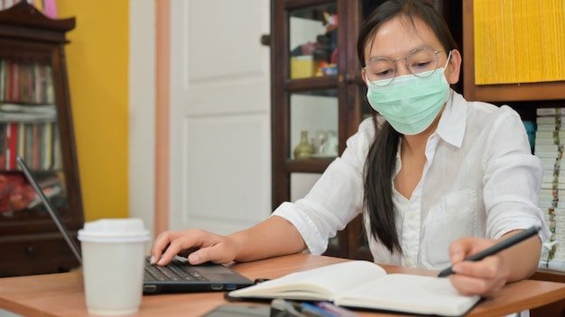 La donna asiatica sta prendendo appunti e utilizzando un computer portatile. lavora a casa per proteggersi dal coronavirus o covid-19.