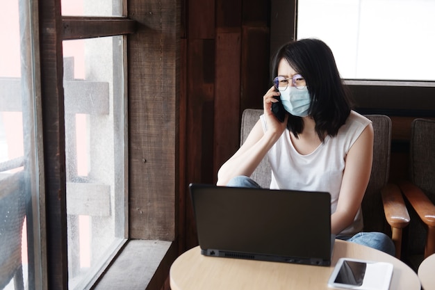 アジアの女性はcovid-19を防ぐためにマスクを着用し、スマートフォンで呼び出しています。実業家は、リビングルームでラップトップを使用してオンラインで作業しています。コロナウイルスの発生の概念のために自宅で仕事をします。