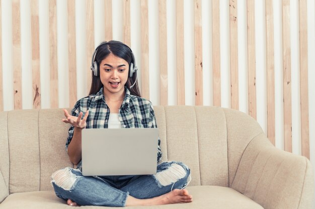 Asian woman in jeans and plaid shirt. Portrait of a young happy female sitting on the couch and working or studying online