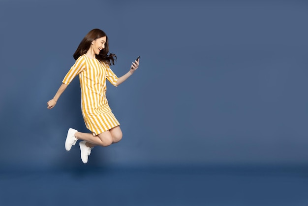 Photo asian woman jumping and using mobile phone application isolated over blue background