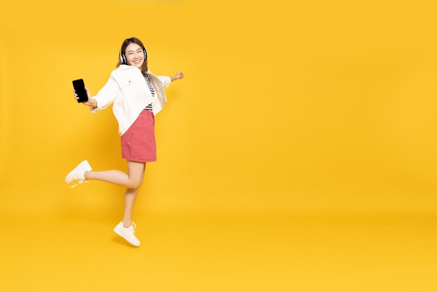 Foto donna asiatica che ascolta la musica preferita sul telefono cellulare e balla isolata su sfondo giallo