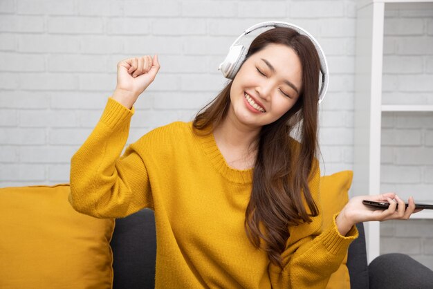 Asian woman listening music with headphones and sitting on sofa in the room at home