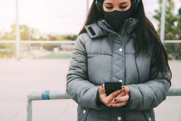 Asian woman looking at cellphone wearing headphones and protective face mask Copy space Lifestyle