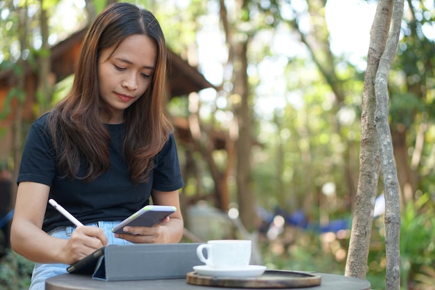 Photo asian woman looking at earnings on her phone at a coffee shop where she can go to work amidst green nature