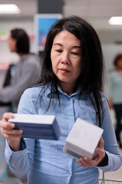 Asian woman looking at supplements packages in pharmacy shop, examining boxes of vitamins and bottles of drugs on shelves. Drugstore customer buying medical products and supplies.