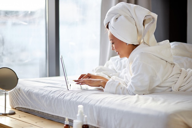 Asian woman lying on bed at home and working on her laptop computer, wearing towel and bathrobe, in the morning. working from home, quarantine coronavirus concept