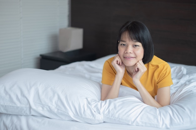 An Asian woman lying on her chin on a white bed in her room