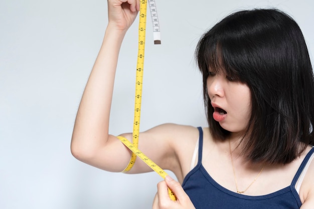 An Asian woman measures her arm with a tape measure and poses in shock at the result