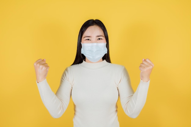 Donna asiatica in maschera medica per proteggere covid19 coronavirus alza le mani felice eccitata allegra su sfondo giallo