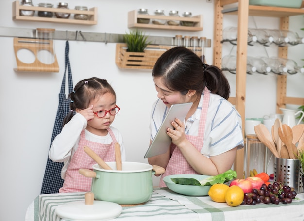 Asian woman mother and daughter play together in kitchen,mom hold tablet for teach little girl how to cook in semester break