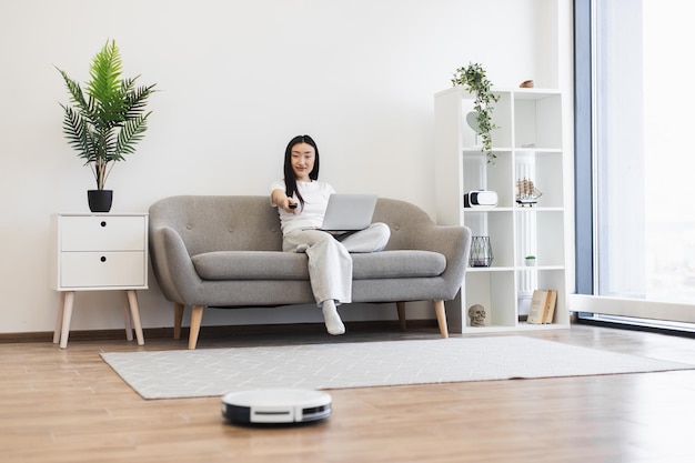 asian woman multitasking with laptop and robot vacuum cleaner at home