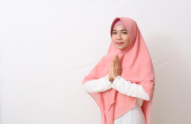 An asian woman in muslim dress stands up in a greetting pose to\
namaste hands, welcoming guests, ied fitr greeting. isolated on\
white background