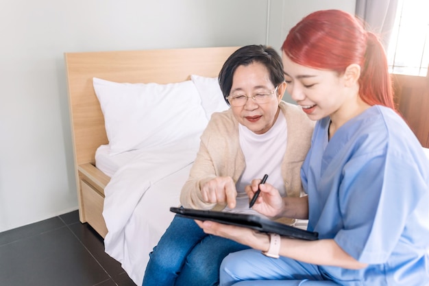 Asian woman nurse wearing scrubs report the health status of Senior Asian woman with digital tablet in the bedroom Caregiver visit at home Home health care and nursing home concept