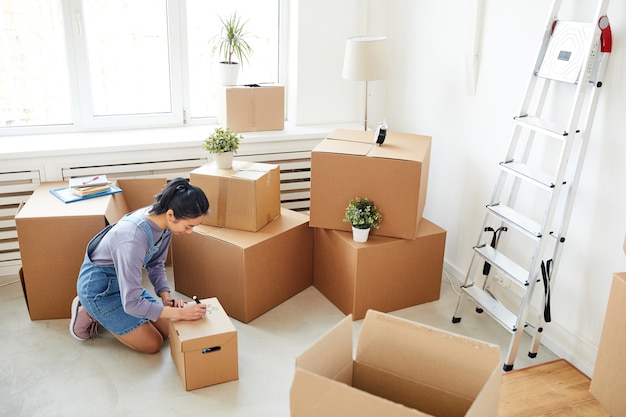 Asian woman packing cardboard boxes in empty white room, moving, relocation and house decor concept