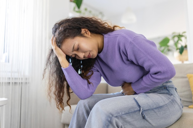 Asian woman in painful expression holding hands against belly suffering menstrual period pain lying sad on home bed having tummy cramp in female health concept