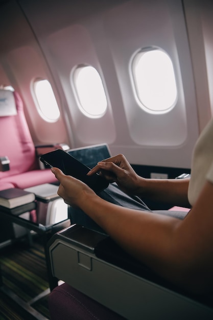 asian woman passenger sitting in airplane near window and reading news from social networks or using travel applications in smartphone
