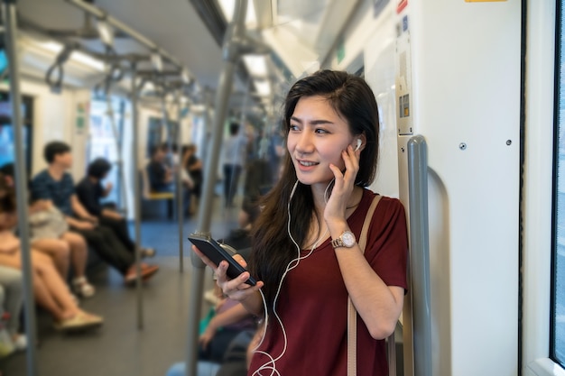 Passeggero asiatico della donna con il vestito casuale che utilizza e che ascolta la canzone tramite il telefono cellulare astuto nel bt