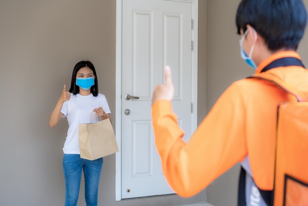 사진 아시아 여자는 문 손잡이에서 배달 음식 가방을 집어 들고 비접촉식으로 엄지 손가락을 올리거나 감염 위험에 대한 사회적 거리를두고 앞 집에 자전거가있는 배달 라이더로부터 무료로 연락하십시오. 코로나 바이러스 개념