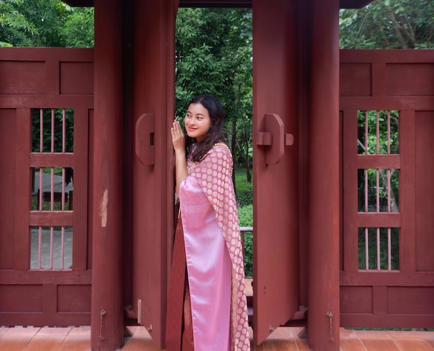 Asian woman in pink Sabai Thai traditional dress peeking at someone from slightly opened old-style wooden door