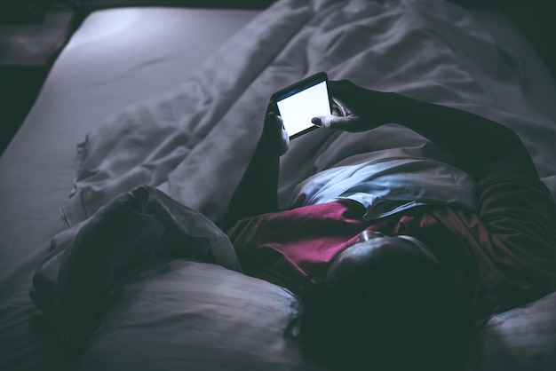 夜にベッドでスマートフォンでゲームをしているアジアの女性タイの人々アディクトソーシャルメディア