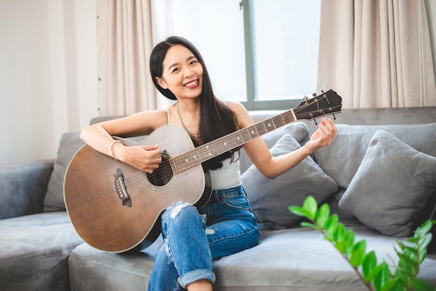 Asian woman playing music by guitar at home, young female guitarist musician lifestyle with acoustic art instrument sitting to play and sing a song making sound in hobby in the house room