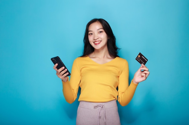 Asian woman posing with a credit card and a phone in the other while standing in front of a light blue background