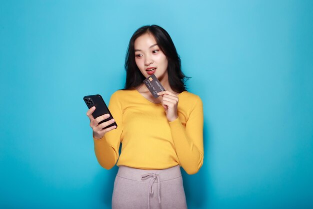 푸른색 배경 앞에 서 있는 아시아 여성이 신용카드와 드폰을 들고 포즈를 취하고 있다.