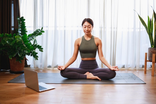 Asian woman practicing yoga from yoga online course on a video\
conference on a laptop at home