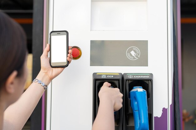 Asian woman preparing her electric vehicle - ev for changing at\
the ev charging station, woman using application on a smartphone to\
connect with ev charging station.