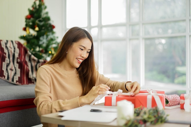 アジアの女性はメリークリスマスのリビングルームで包まれたギフトボックスにリボンを置きました