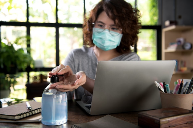 Asian woman in quarantine and social distancing wearing surgical mask and cleaning hands with alcohol gel sanitizer while working from home during Covid-19 Coronavirus pandemic