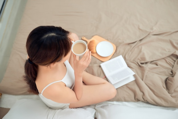아침에 침대에서 커피 한 잔과 함께 책을 읽는 아시아 여성