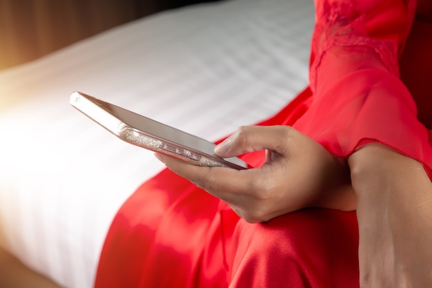 Asian woman in red satin robes using a mobile phone while sitting on a bed at night