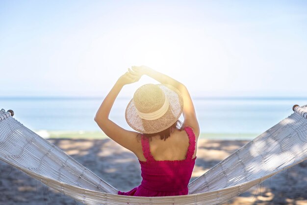 Photo asian woman relaxing in the hammock on tropical beach njoy her freedom and fresh air wearing stylish hat and clothes happy smiling tourist in tropics in travel vacation