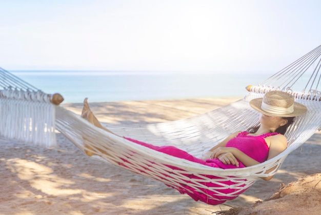 Photo asian woman relaxing in the hammock on tropical beach njoy her freedom and fresh air wearing stylish hat and clothes happy smiling tourist in tropics in travel vacation