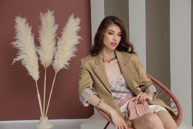 Photo asian woman in sandy brown jacket blazer cream skirt top necklace bracelets rosy brown wall