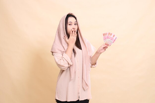 Photo asian woman shocked at the camera hands covering her mouth and carrying rupiah money young beautiful