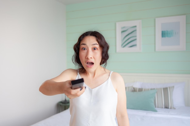 Азиатская женщина в шоке и чувствует себя ВАУ, когда включает телевизор с помощью пульта дистанционного управления Азиатская домохозяйка проверяет социальные сети с помощью смарт-телевизора дома OMG